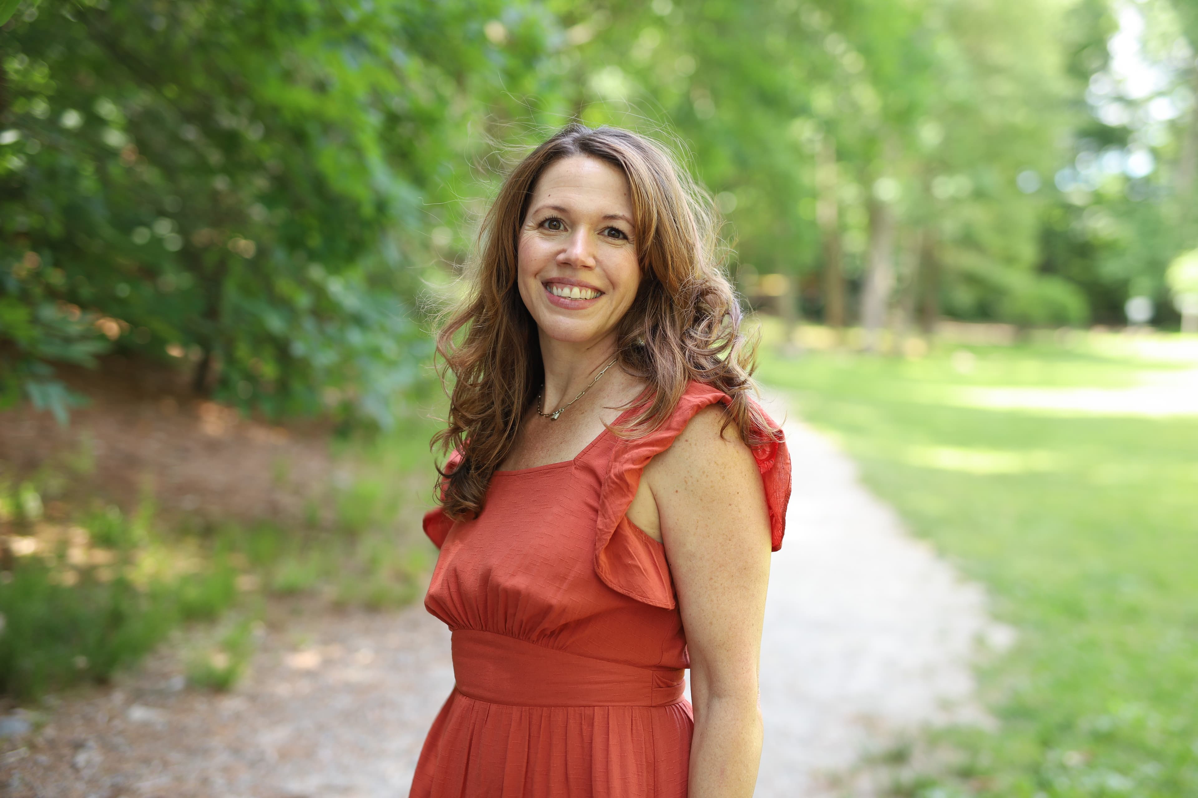 Woman smiling in a red dress standing on a park path with trees in the background.