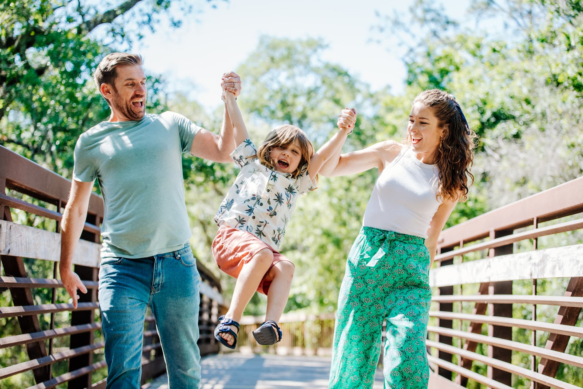 A happy family swinging a child by the arms on a sunny walkway.