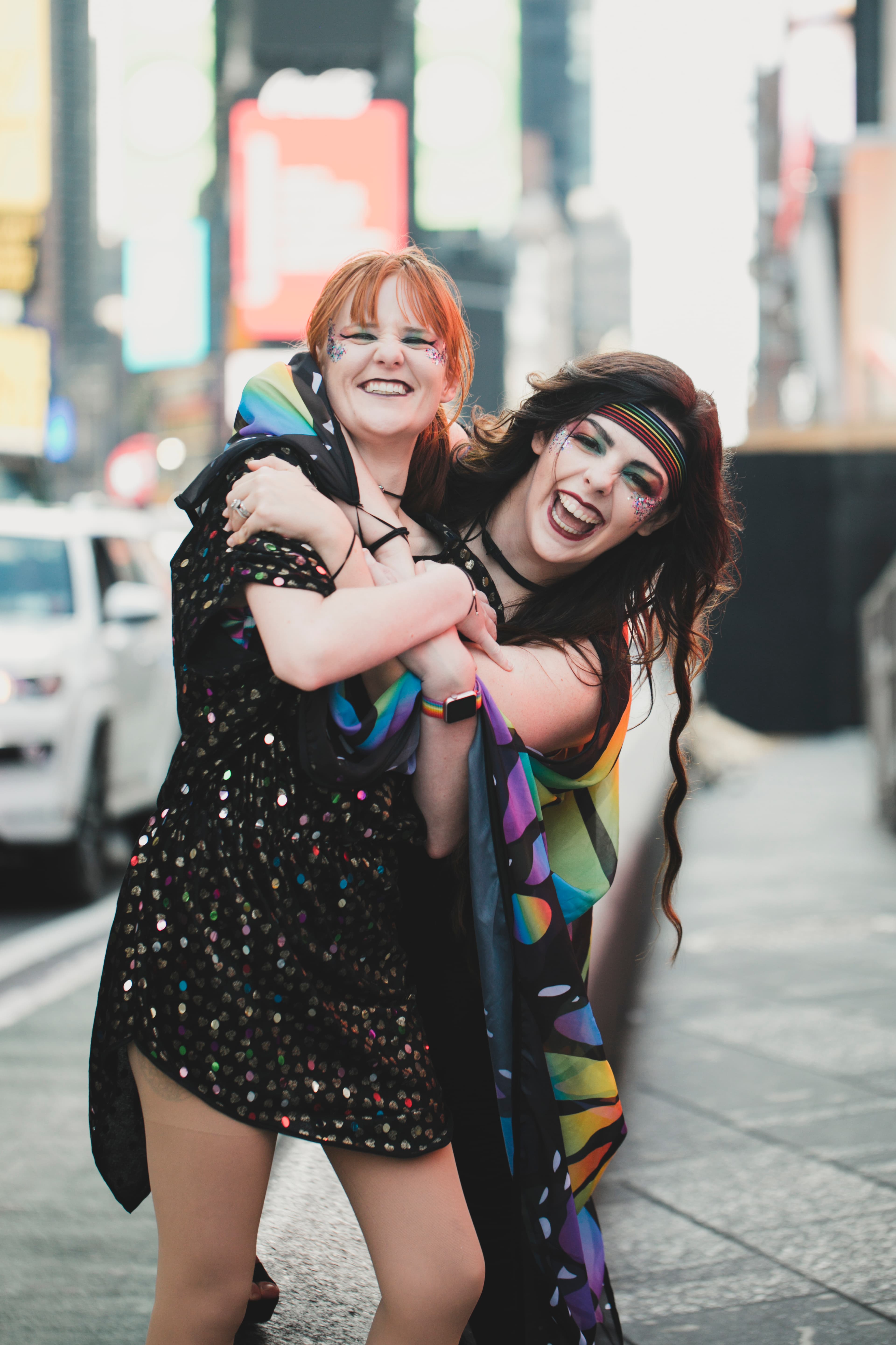 Two joyful people hugging and smiling on a city street with bright makeup and colorful outfits.