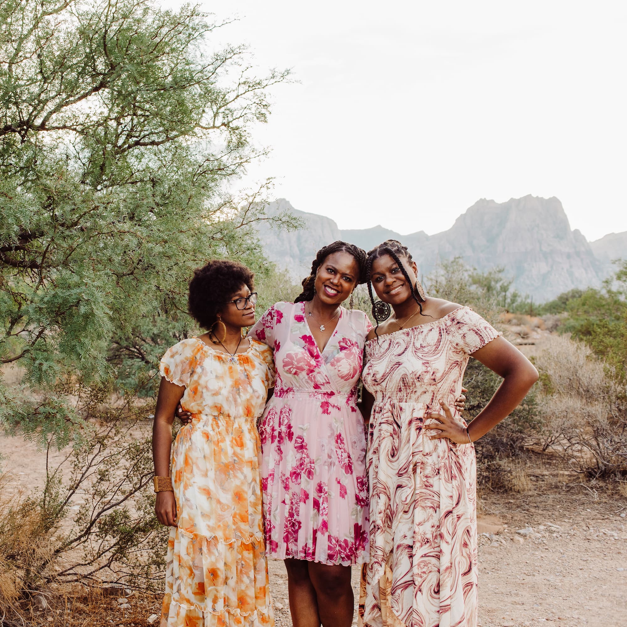 Three women smiling in colorful dresses standing in a desert landscape.