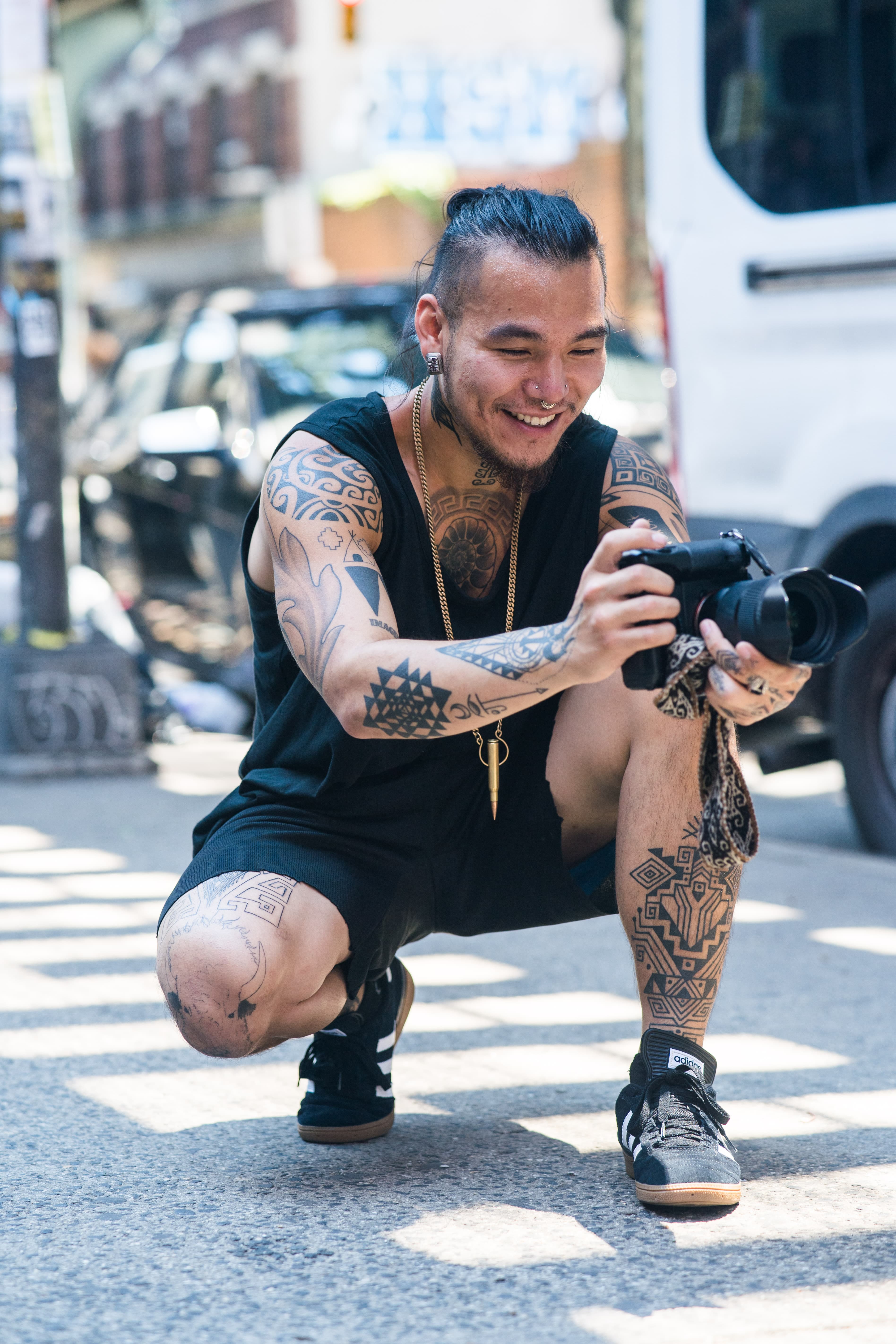 A tattooed photographer crouches with his camera on a city street.