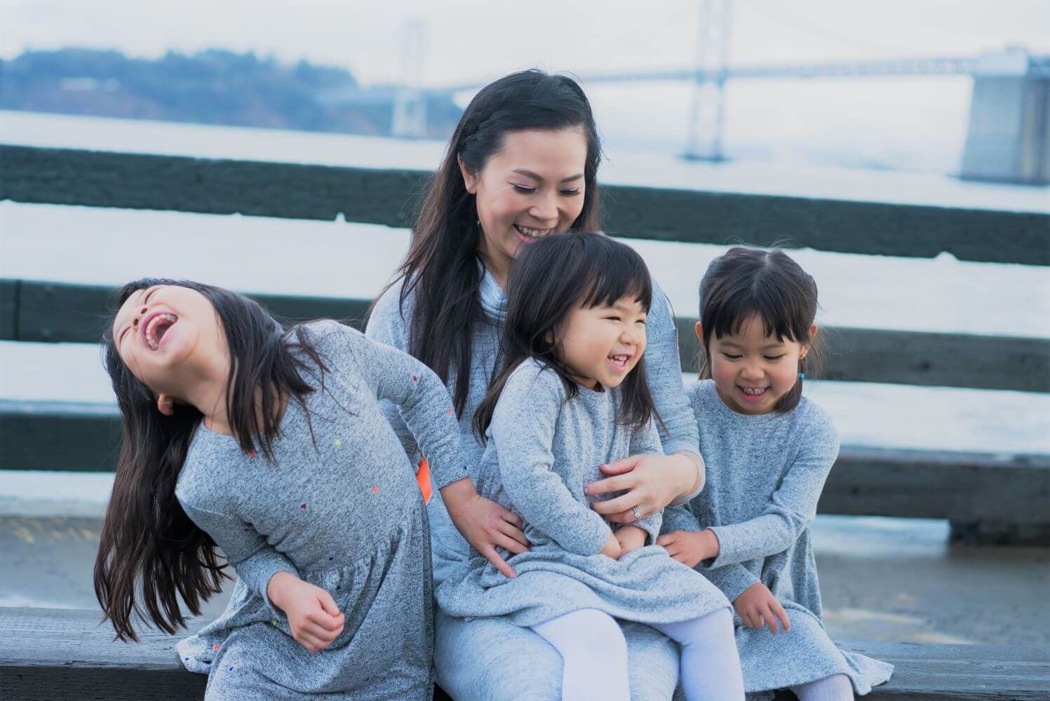 Three young girls and a woman are laughing together while sitting on a bench with a bridge in the background.