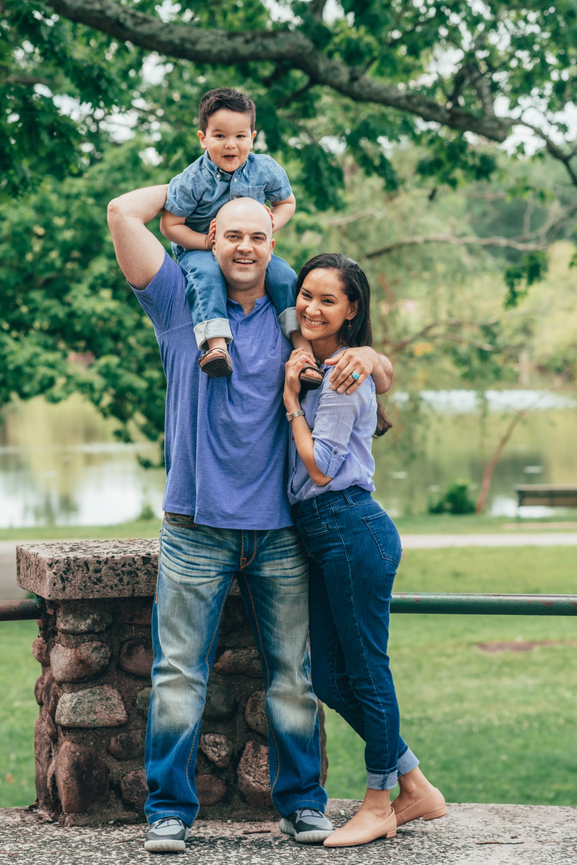 A family of three, with a child on his father's shoulders, smiling at a park.
