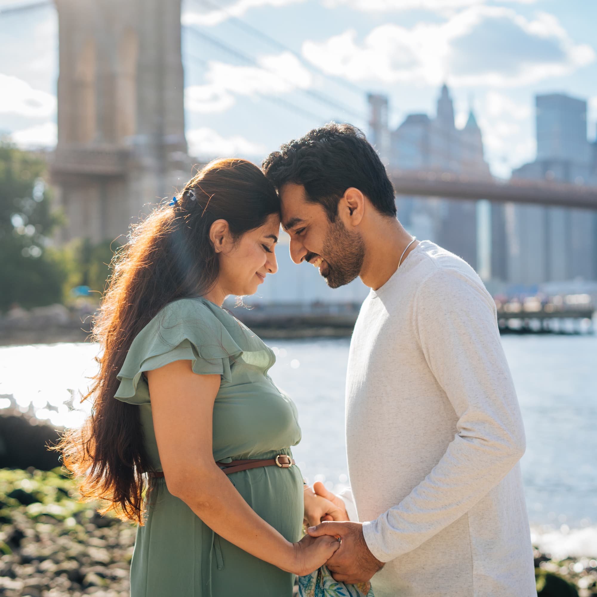 A smiling couple with the man's hand on the woman's pregnant belly, with a bridge in the background.