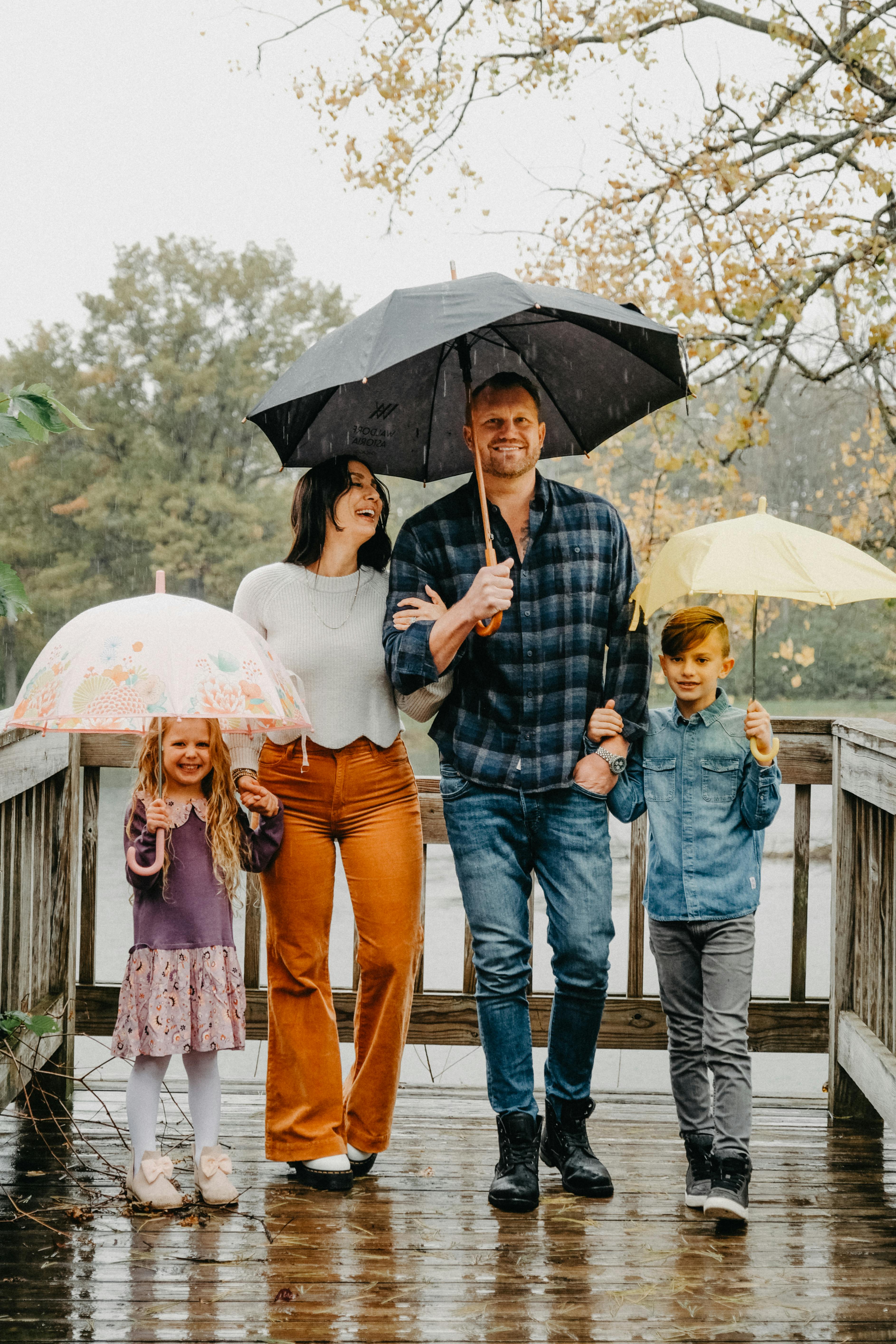 A family of four with umbrellas smiling and walking on a wooden bridge in the rain.