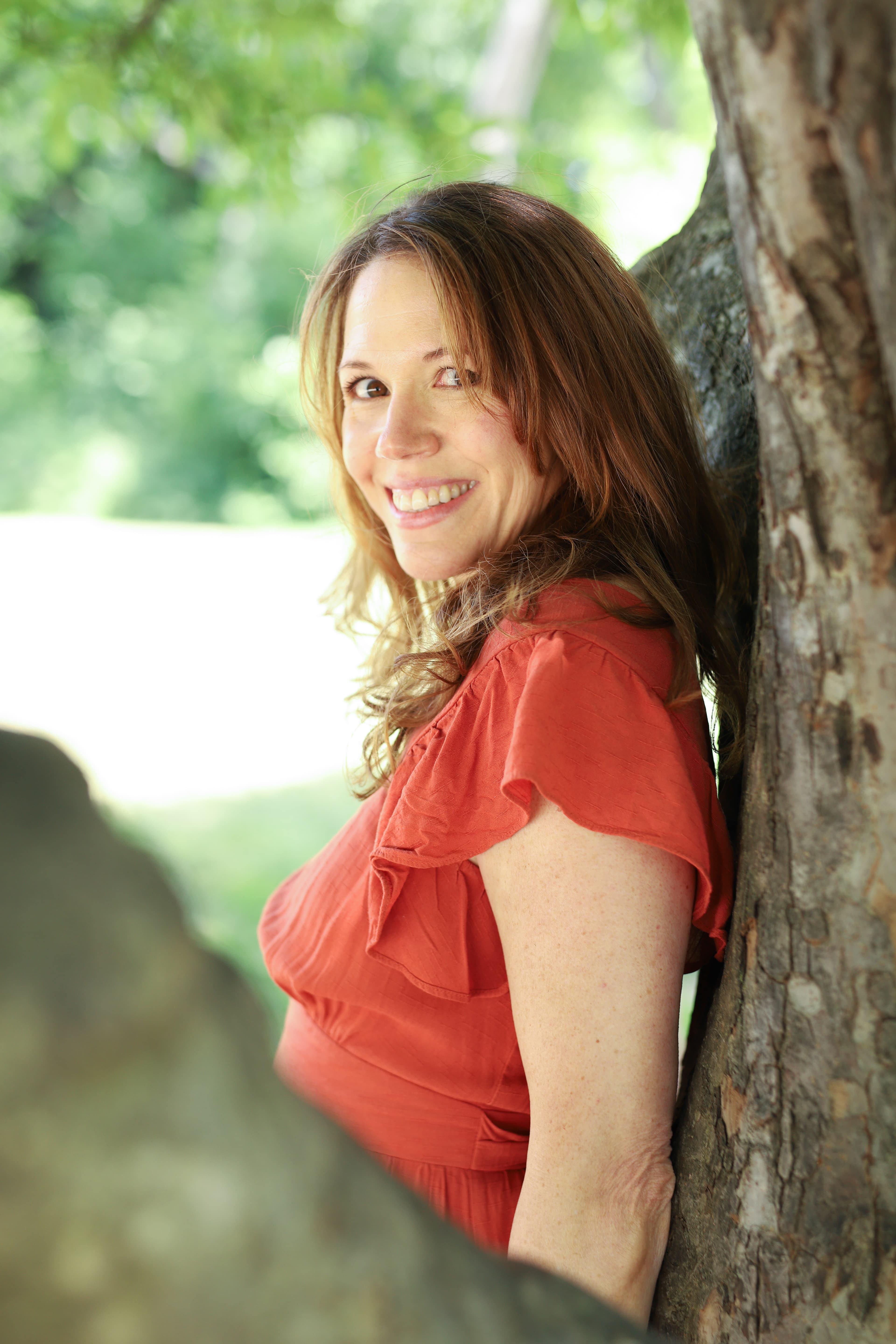 Smiling woman in a red dress leaning against a tree.