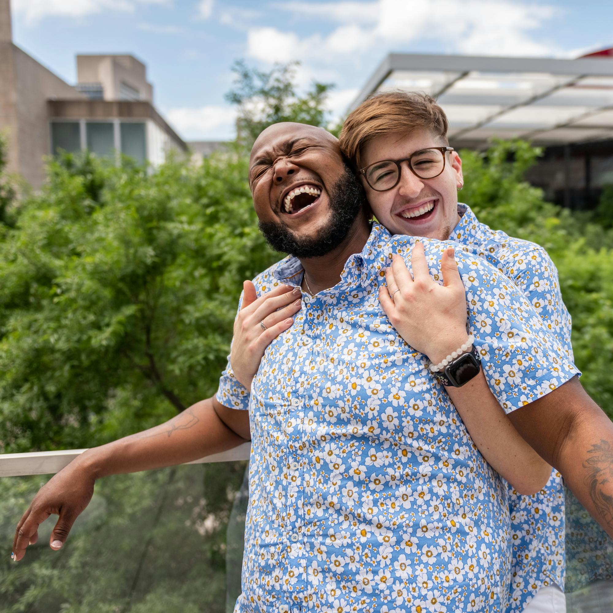 Two people laughing and embracing in a friendly gesture, both wearing similar floral shirts.
