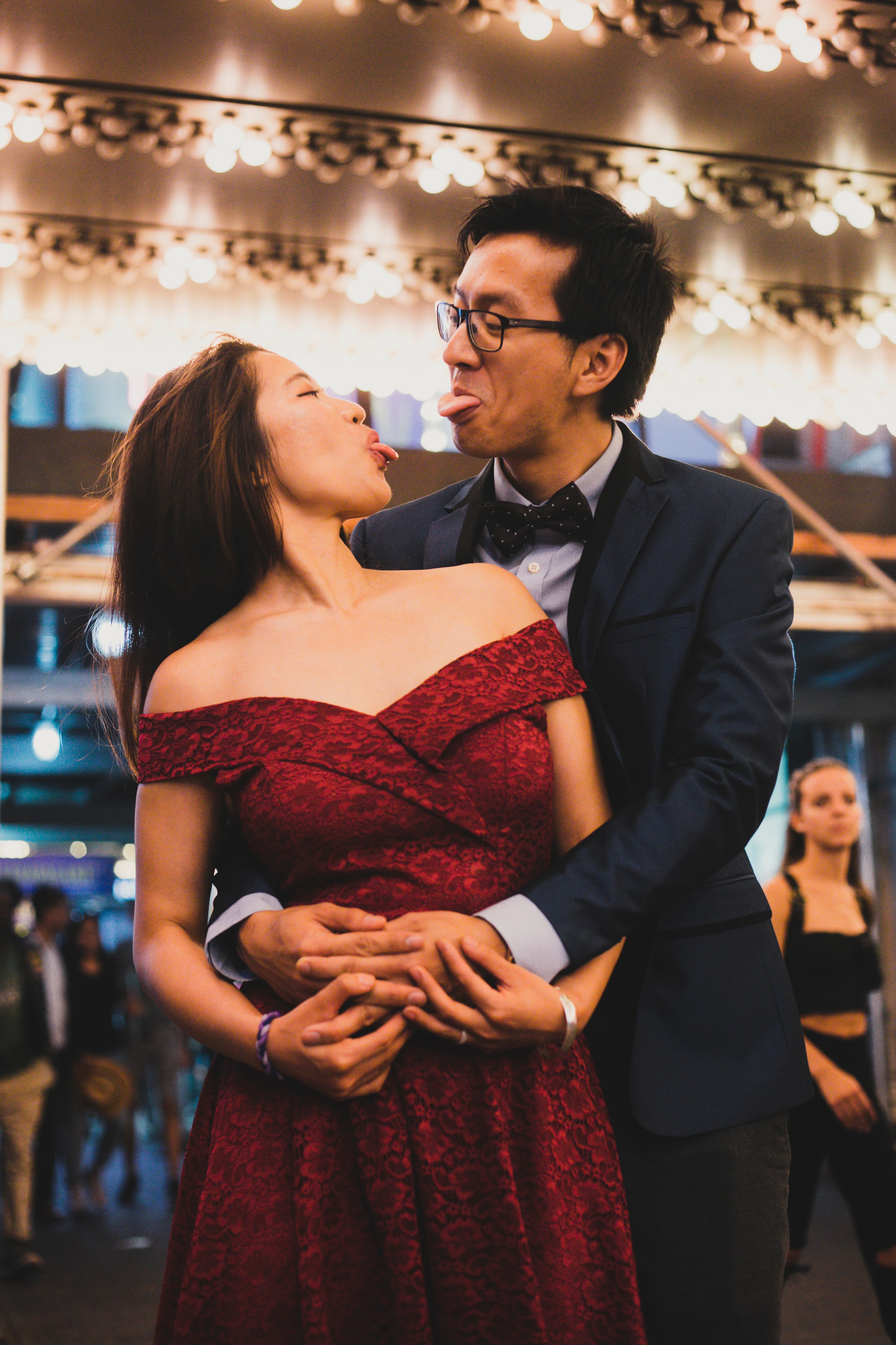 A couple in formal attire making playful faces at each other under festive lights.