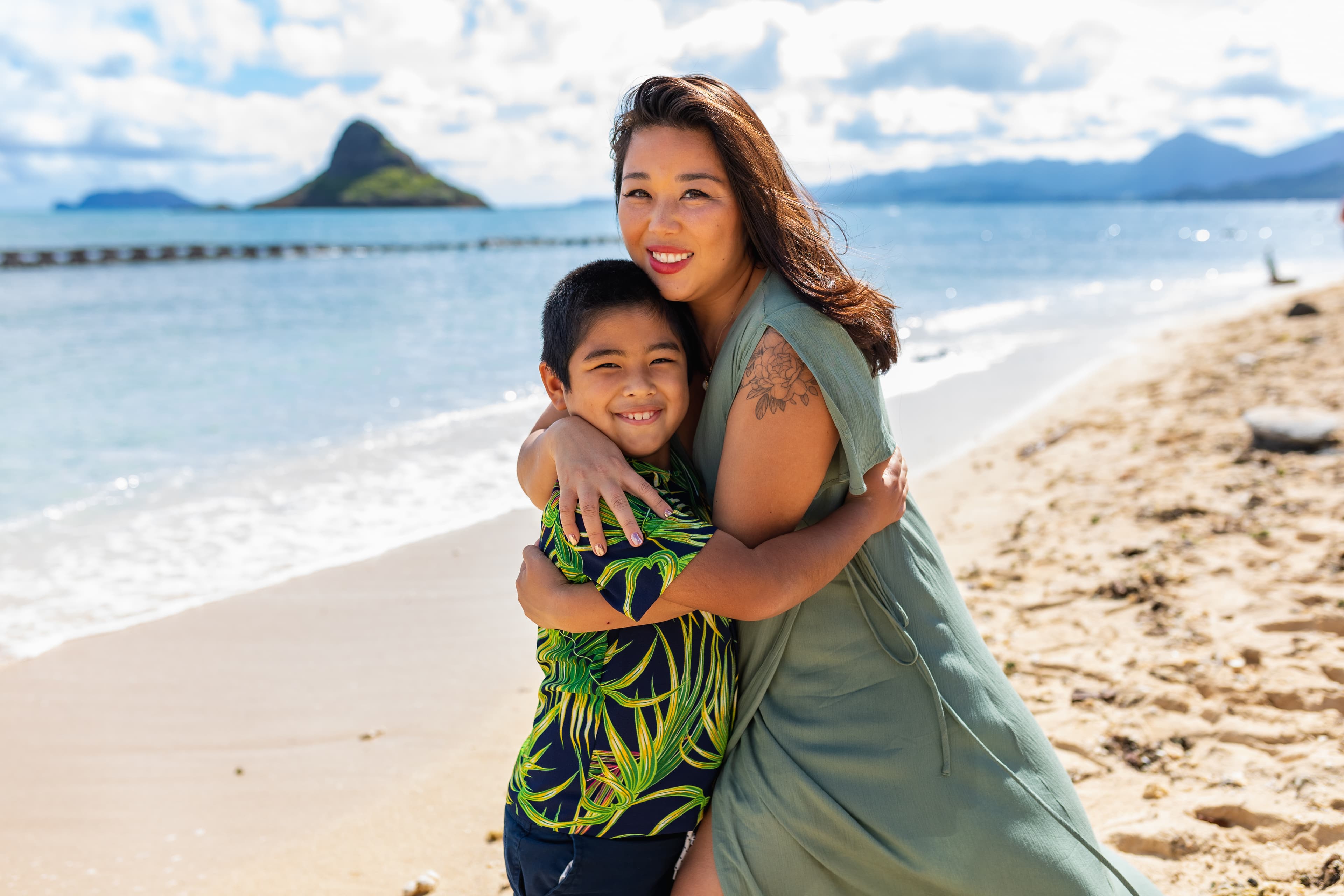 A smiling woman embracing a young boy on a sunny beach with a mountain in the background.