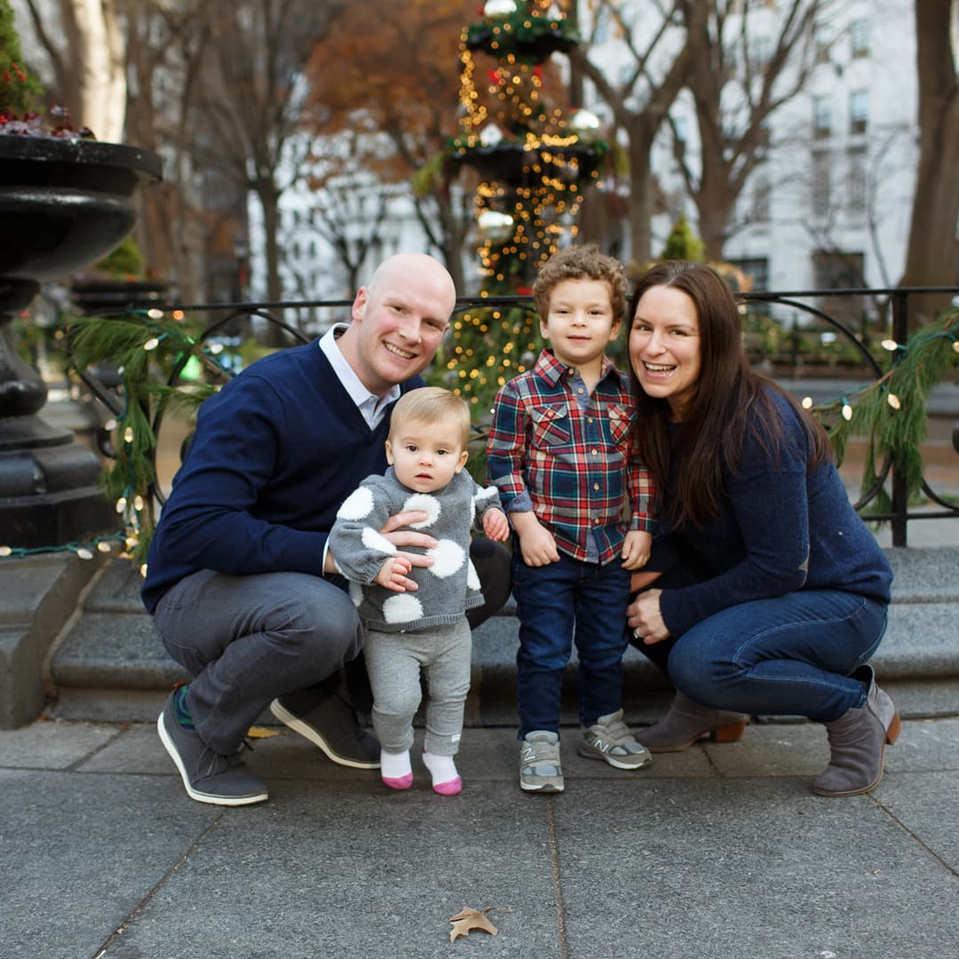A family of four crouches together for a photo in front of a city park fountain with holiday decorations.