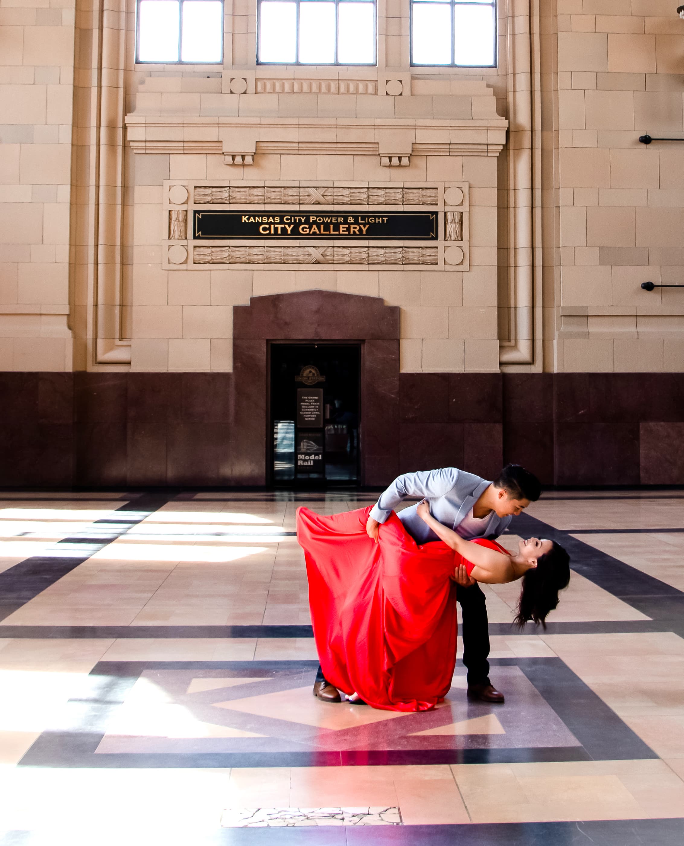 A couple performing a dance move in a grand hall with the woman wearing a red dress and the man in a gray suit.