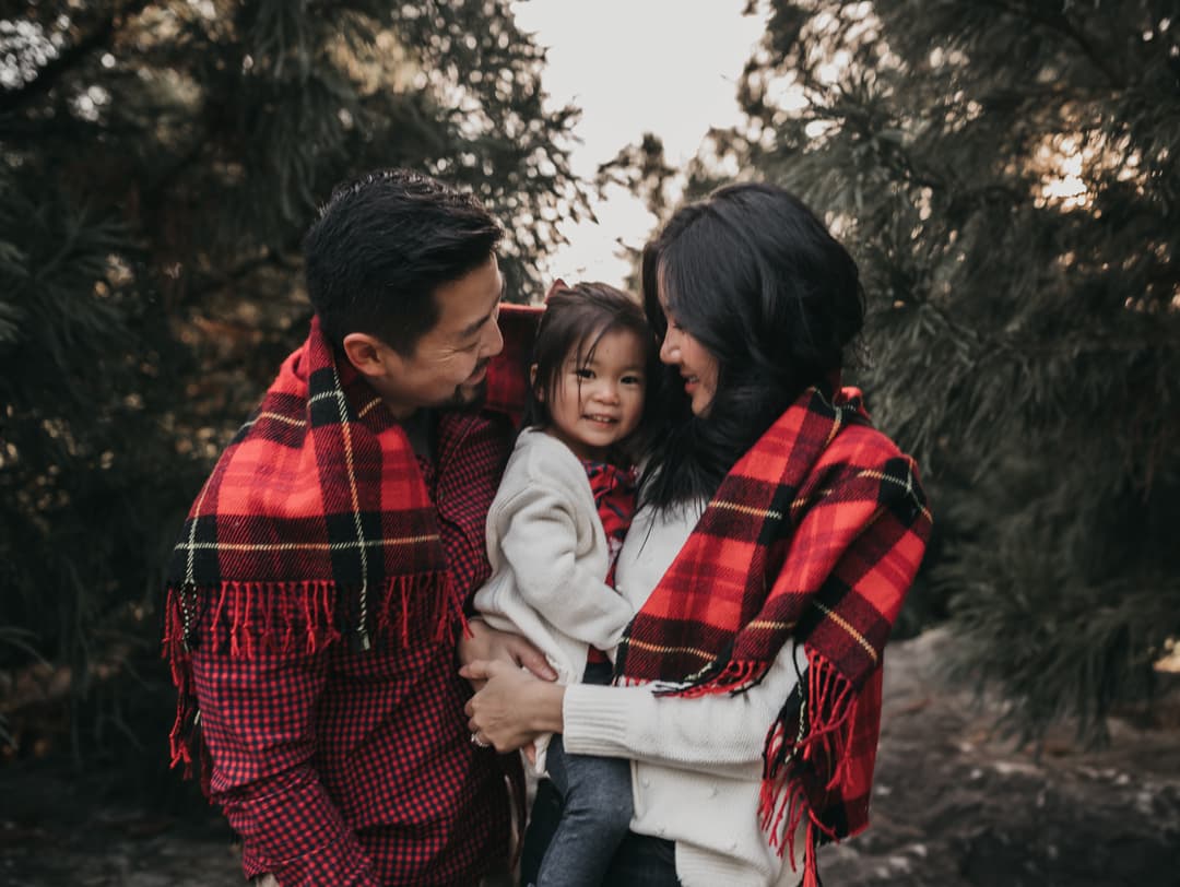 A happy family with a child wrapped in a red plaid blanket standing in a forest.