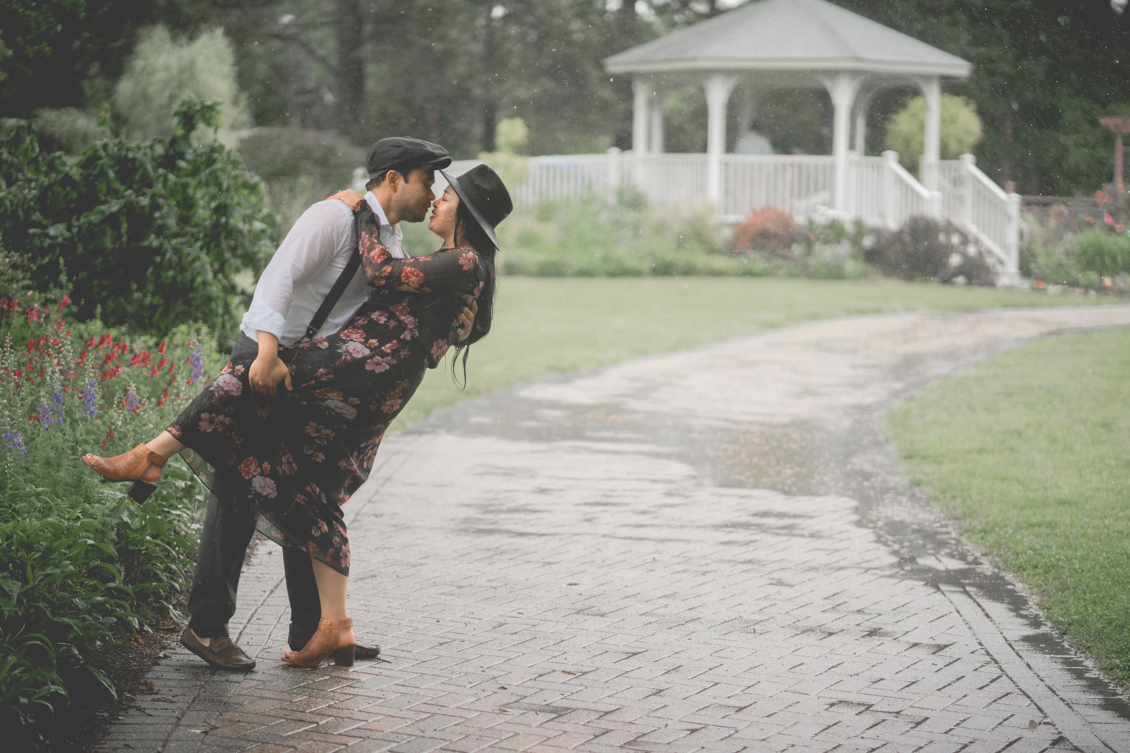 A couple sharing a romantic kiss in the rain on a park path near a gazebo.