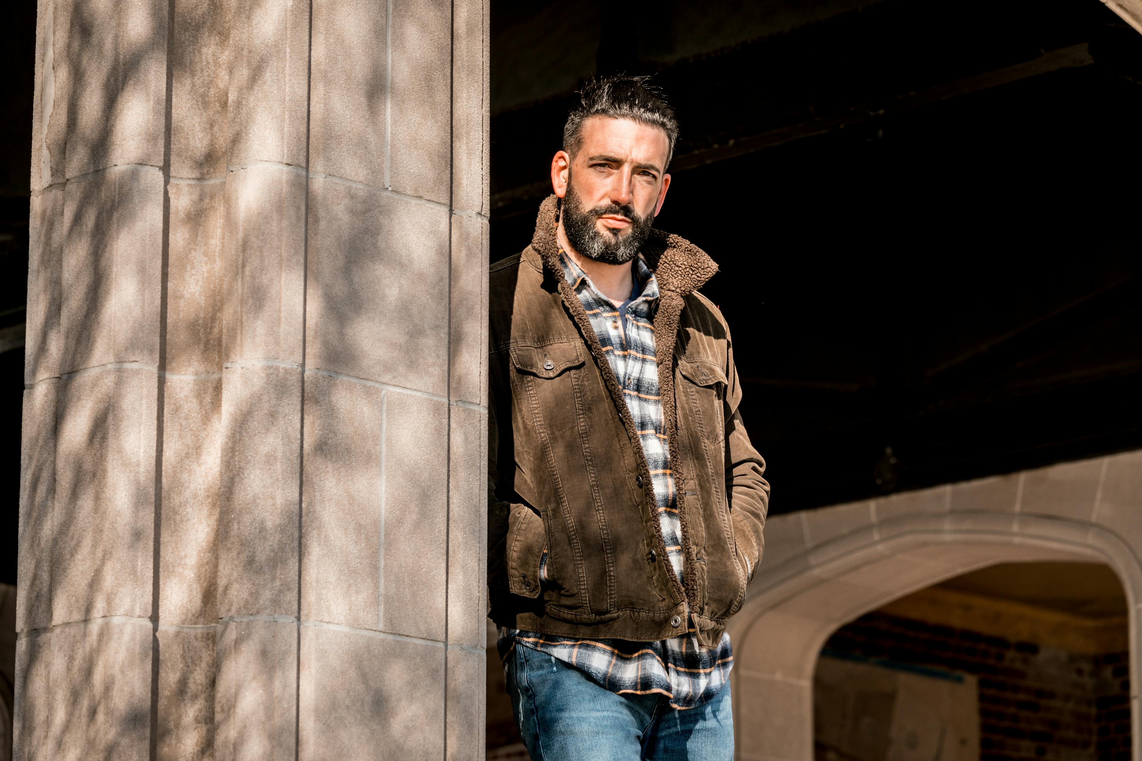 Man leaning against a column with a thoughtful expression in a casual jacket and plaid shirt.