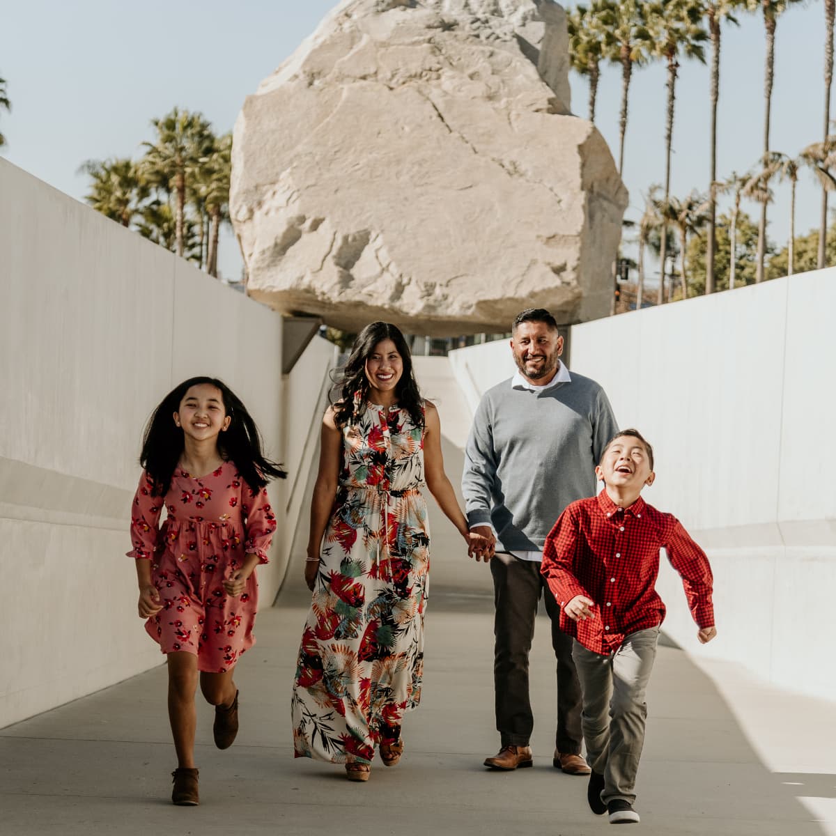 Family of four walking hand in hand and smiling under a suspended boulder installation with palm trees in the background.