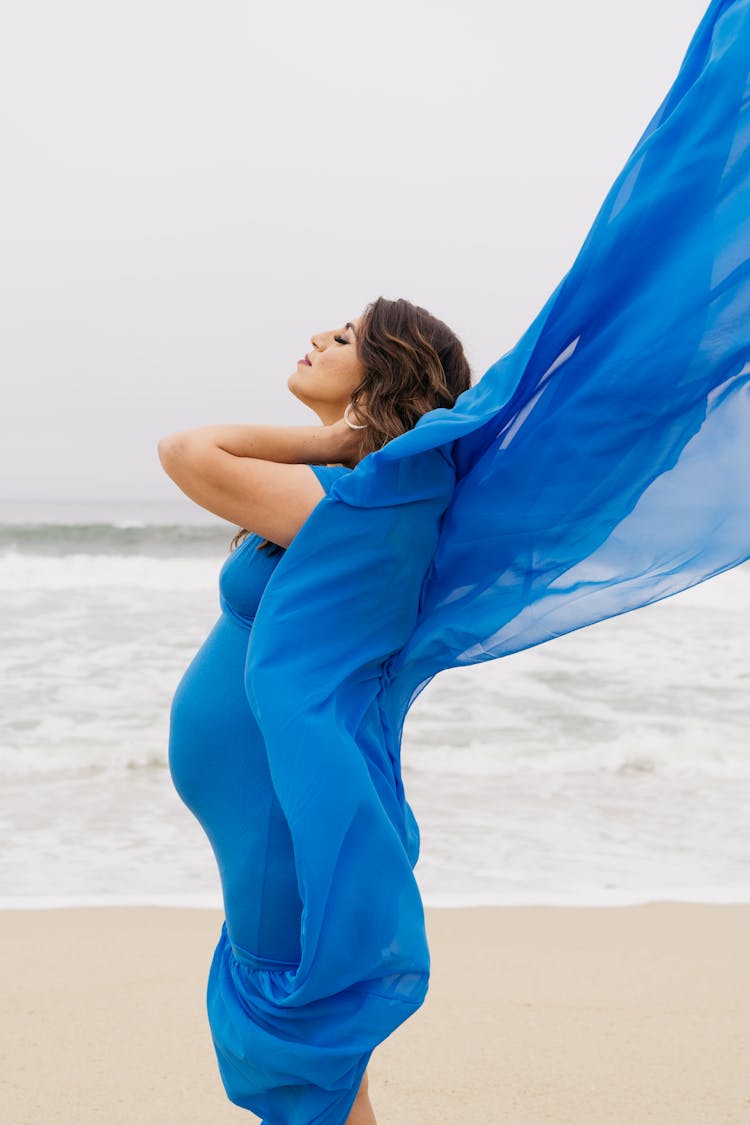 Woman standing on beach in long, flowy, blue maternity gown.