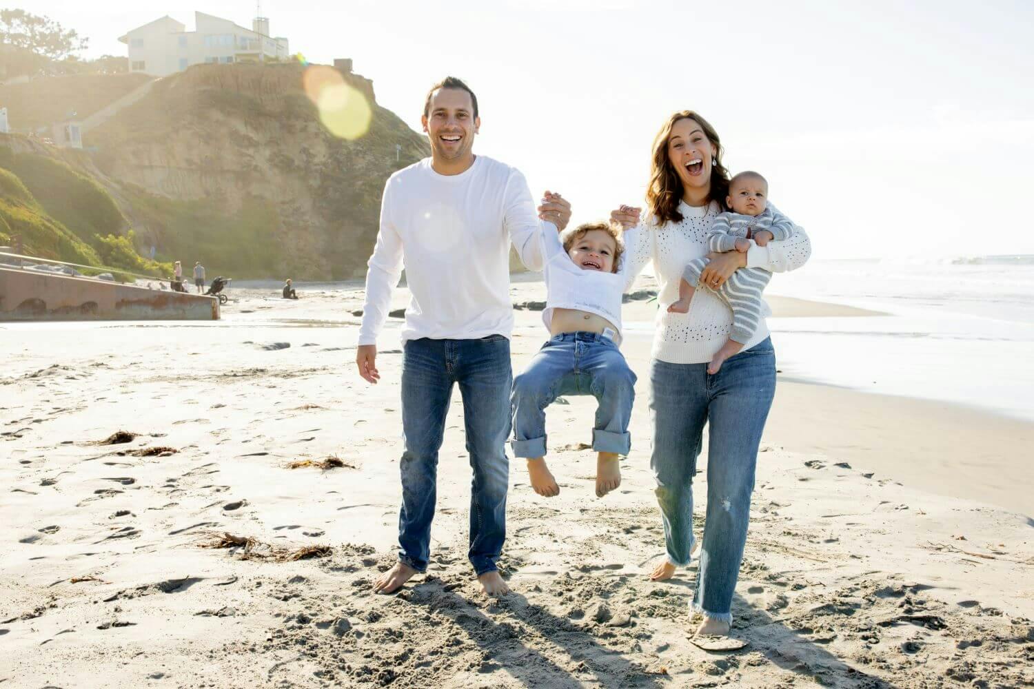 A family of four laughing and walking on the beach, with one child being held in the air.