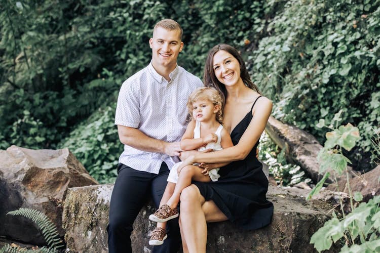 Family of three sitting and smiling on a rock in a lush outdoor setting