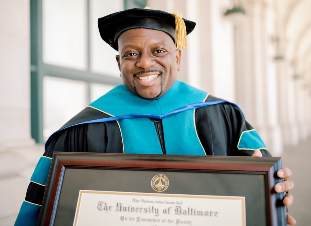 Graduate in cap and gown smiling and holding a framed diploma during a Shoott graduation photoshoot.