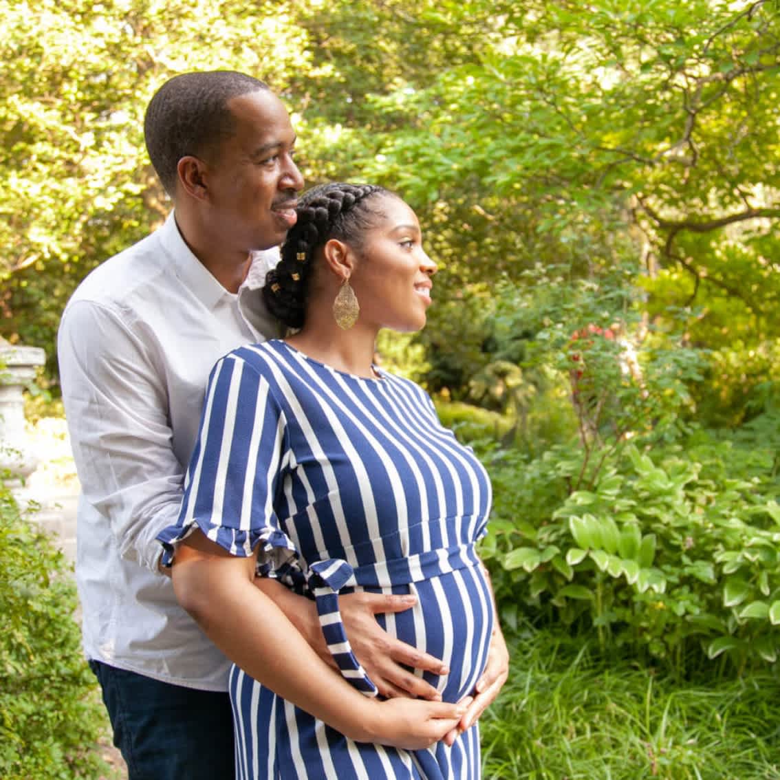 Expectant couple embracing outdoors with the man standing behind the woman, who is cradling her baby bump.