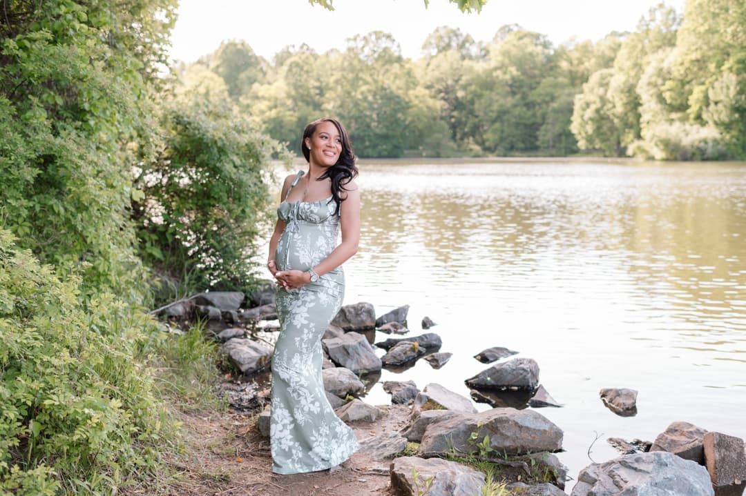 Pregnant woman in a green floral dress standing by a lakeside, smiling and holding her baby bump, surrounded by trees and rocks in a natural outdoor setting