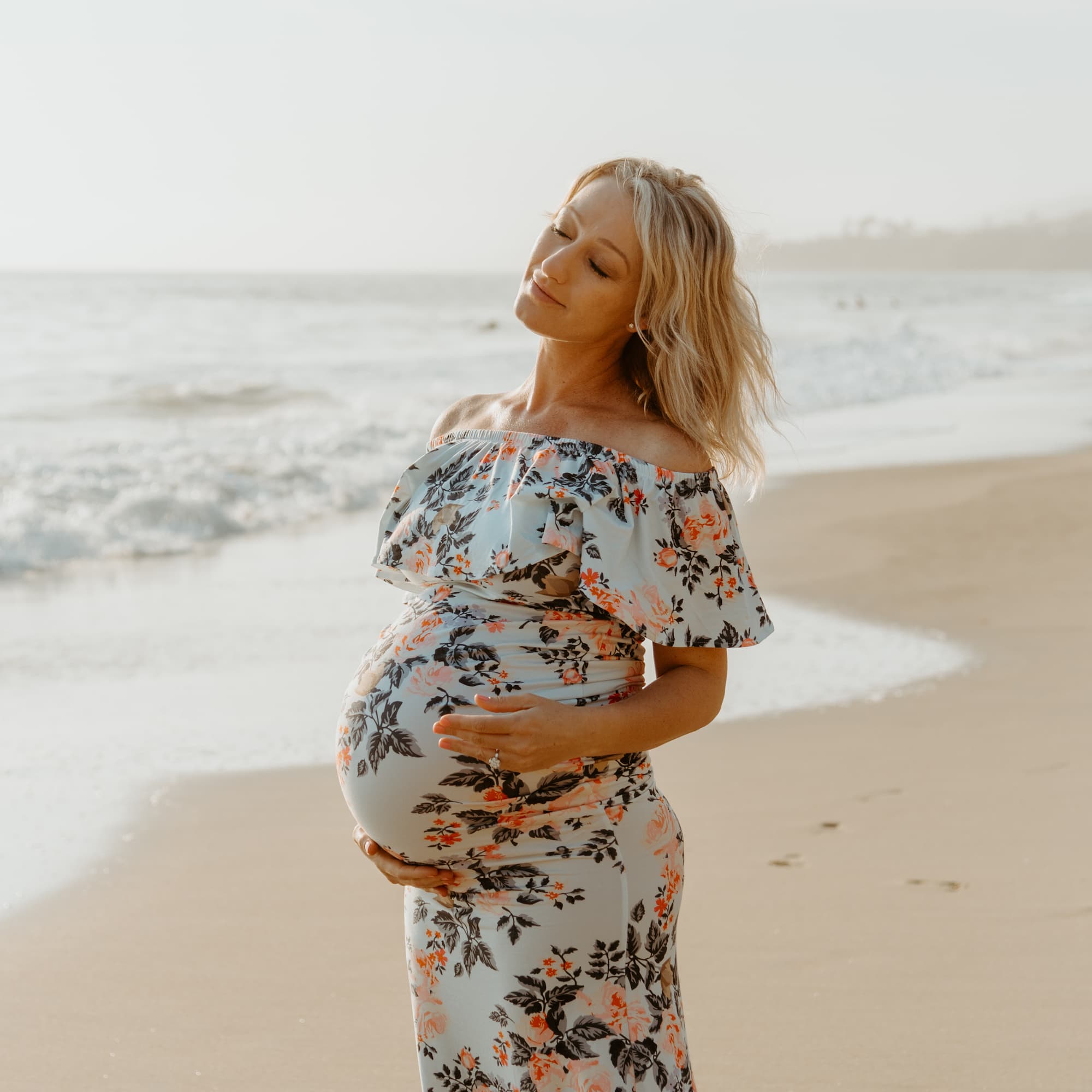 Pregnant woman in a floral dress standing on the beach with her eyes closed.