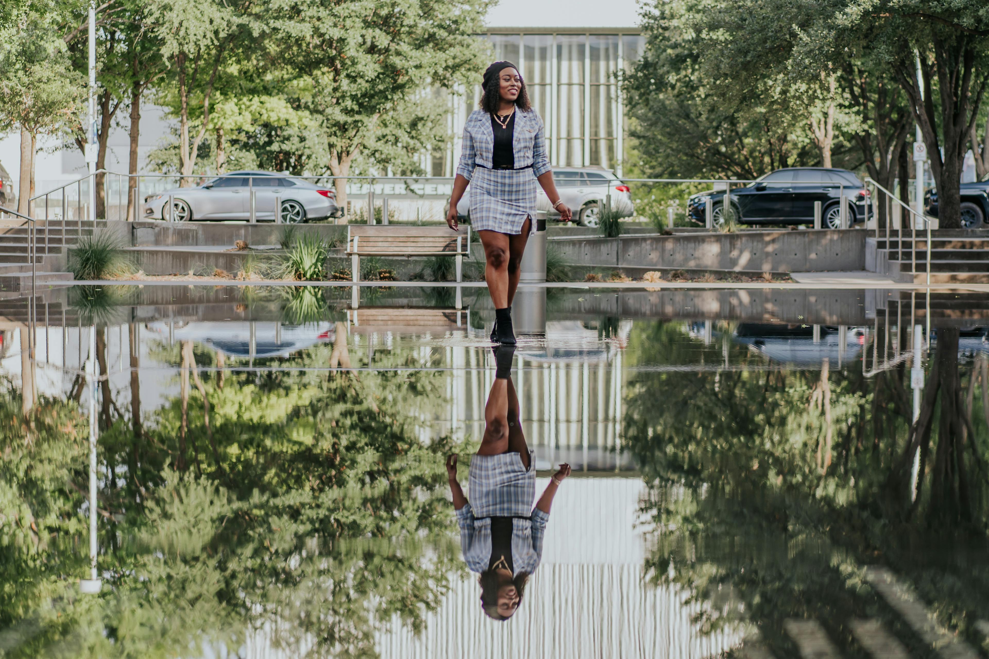 A woman smiling as she walks across a shallow reflecting pool with trees and cars in the background.