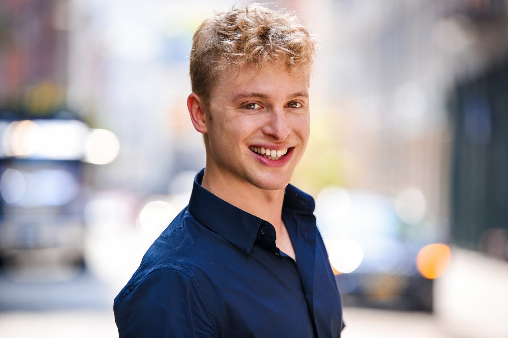 Smiling young man with blond hair in a blue shirt on a city street.