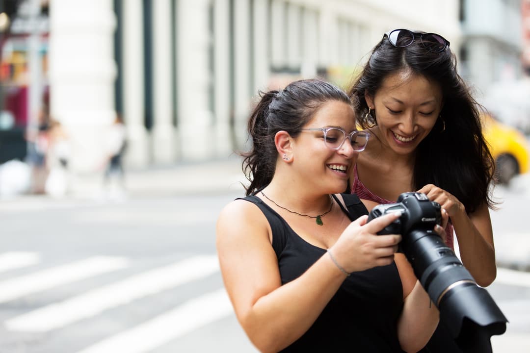 Two women smiling and looking at a photo on a digital camera in an urban setting.